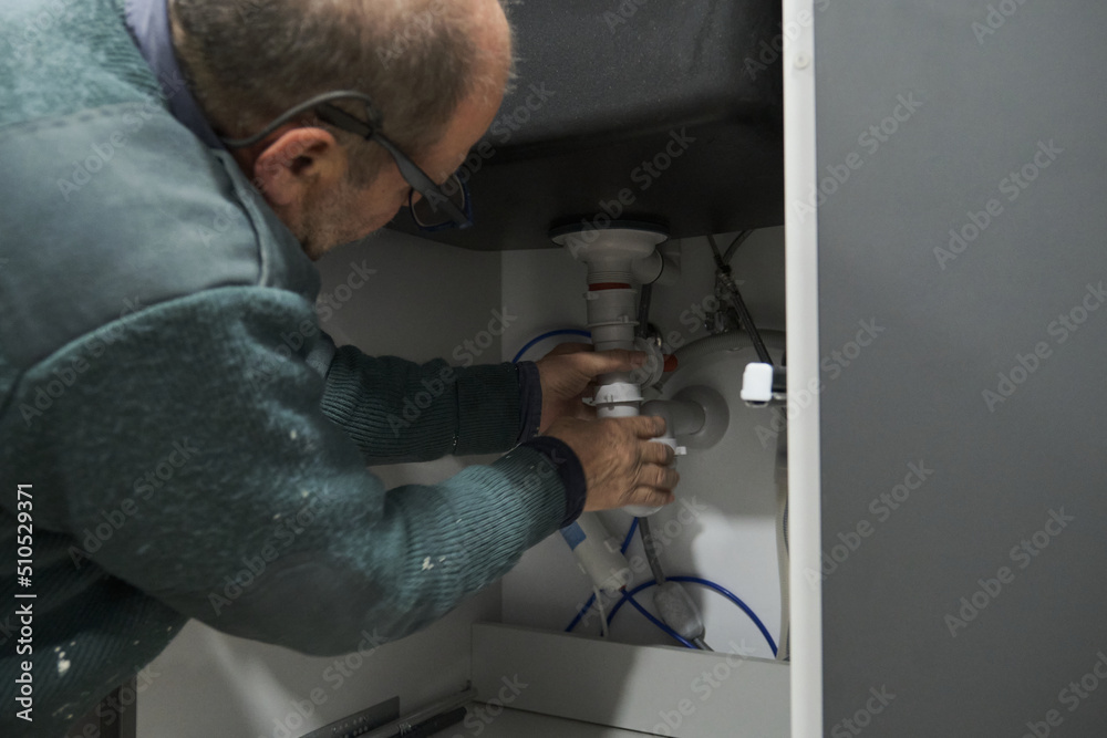 Mature plumber installing sink siphon in domestic kitchen. Repairman working.