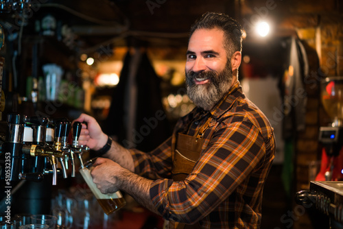 Portrait of cheerful barmen at pub. He pours beer into a beer glass.