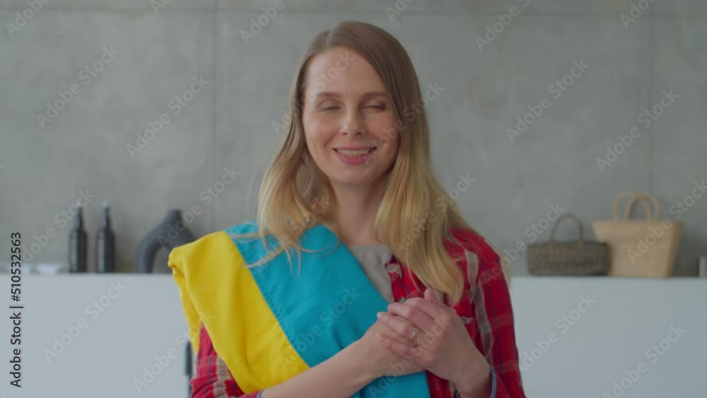 Portrait of charming middle aged woman with flag of Ukraine over shoulder holding hands on heart, looking with friendly smile, expressing pride, national identity and patriotism in home interior. Portrait of charming middle aged woman with flag of Ukraine over shoulder holding hands on heart, looking with friendly smile, expressing pride, national identity and patriotism in home interior.
