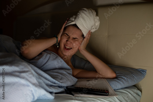 Young woman with white towel wrapped around head lying on bed suffering from headache, earache covering ears with hands. photo