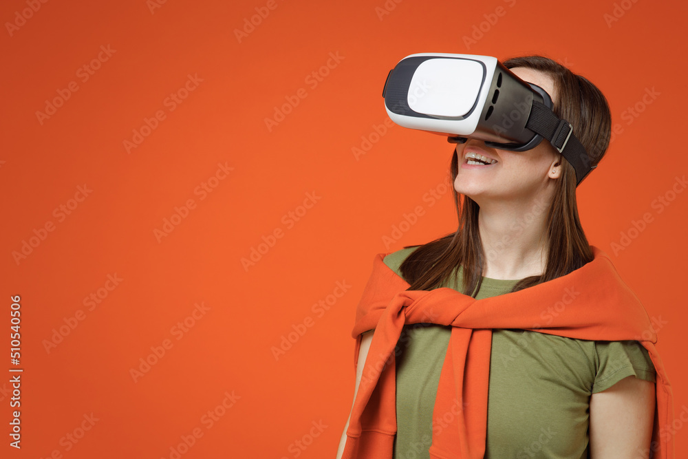 Young amazed happy woman 20s wearing khaki t-shirt tied sweater on shoulders look aside watching in vr headset pc gadget isolated on plain orange background studio portrait. People lifestyle concept. Young amazed happy woman 20s wearing khaki t-shirt tied sweater on shoulders look aside watching in vr headset pc gadget isolated on plain orange background studio portrait. People lifestyle concept.