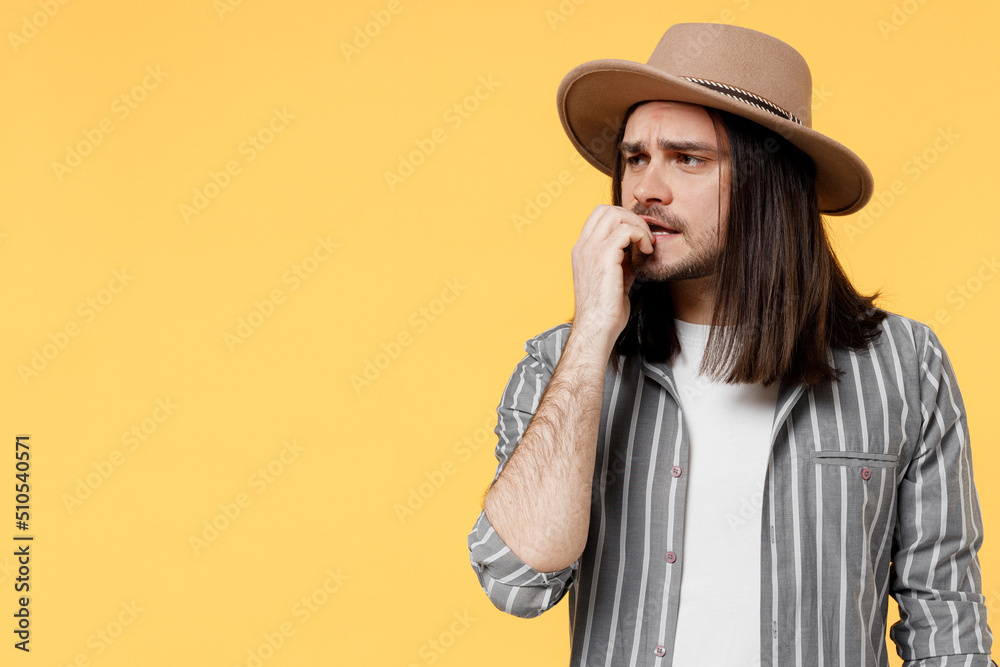 Young confused man he 20s wears striped grey shirt white t-shirt hat look aside on workspace biting nails fingers isolated on plain yellow color background studio portrait. People lifestyle concept. Young confused man he 20s wears striped grey shirt white t-shirt hat look aside on workspace biting nails fingers isolated on plain yellow color background studio portrait. People lifestyle concept.