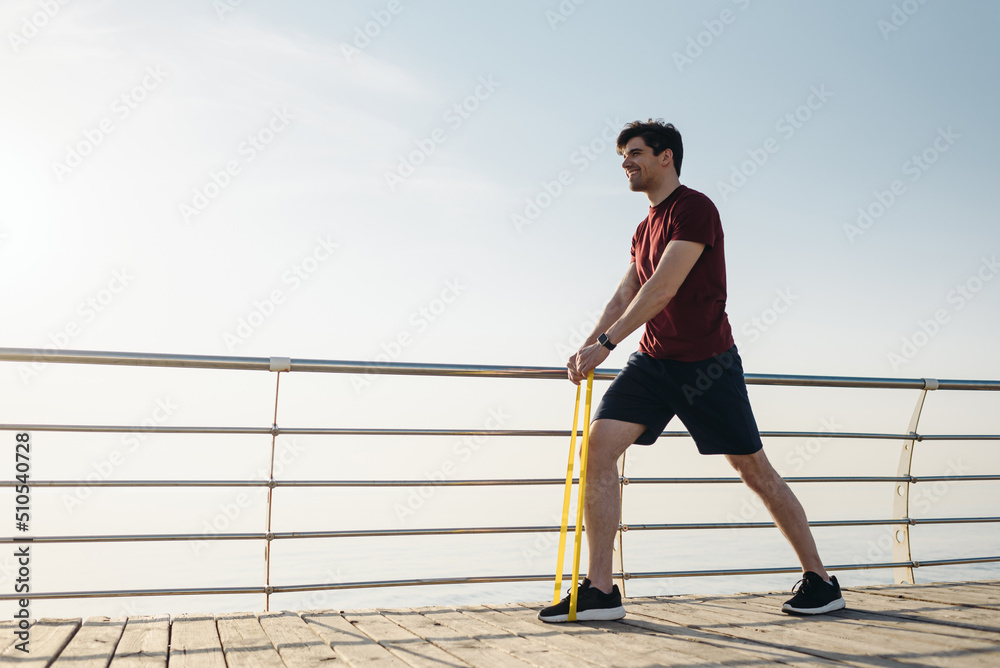 Full size side view young strong sporty athletic toned fit sportsman man wear sports clothes resistance bands warm up training at sunrise sun dawn over sea beach outdoor on pier seaside in morning