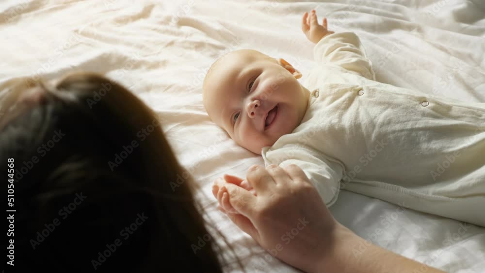 Happy smiling little baby girl lies on white sheet in bright room and enjoys mom attention. Mother hand gently holds newborn daughter tiny hand