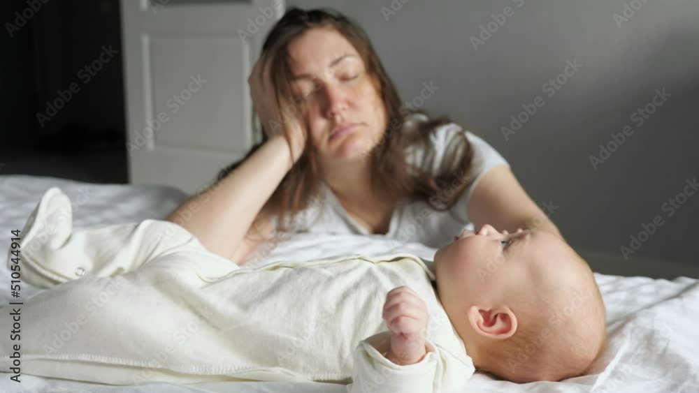 Tired mother sleeps sitting on floor leaning on bed with crying baby lying on bed white sheets. Newborn girl tries to draw attention of mother by crying Tired mother sleeps sitting on floor leaning on bed with crying baby lying on bed white sheets. Newborn girl tries to draw attention of mother by crying