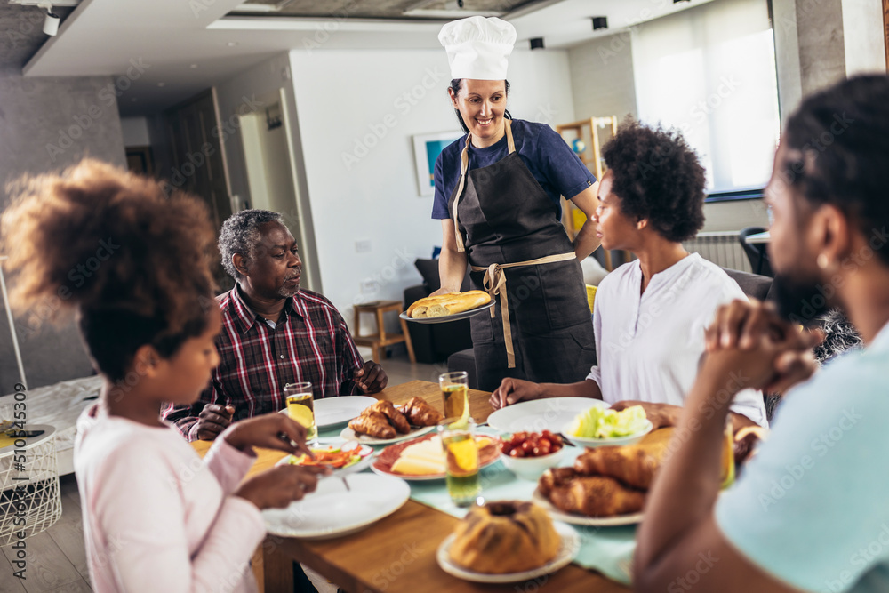 A multi-generational African-American family enjoying food at their dinner table. Caucasian woman cook serve them.