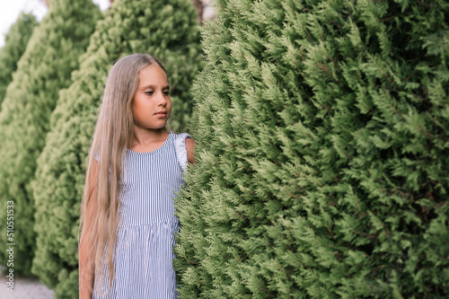 candid caucasian happy little kid girl of eight years old with long blond hair and green eyes on background of green plants during a summer vacation travel. gen z mental health concept © Ksenia