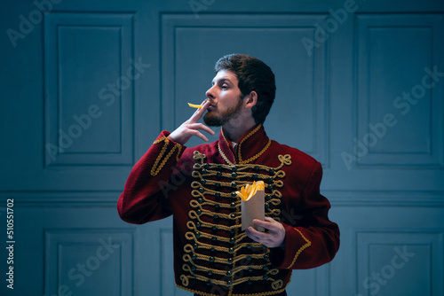 Photos Comic portrait of young man in image of medieval hussar smoking fries isolated on dark blue background