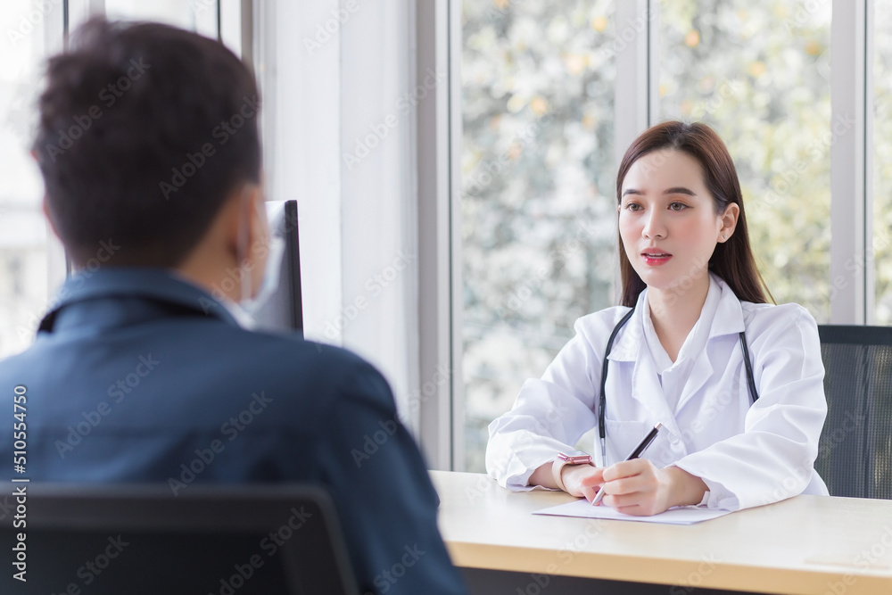 Asian professional  woman doctor who wears medical coat talks with a man patient to consult and suggest healthcare information to him at the examination room in the hospital.