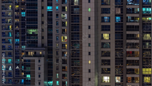 Apartment windows of a glazed skyscraper glow at night with city lights reflection aerial timelapse.