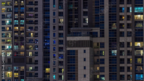 Apartment windows of a glazed skyscraper glow at night with city lights reflection aerial timelapse.