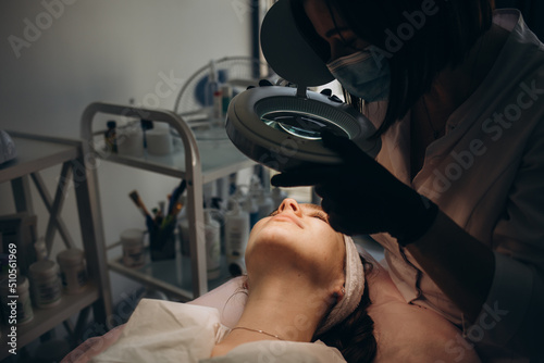 A beautician wearing medical gloves cleans the pores on the face using special equipment while looking through a magnifying glass. The concept of facial hygiene, cosmetic procedures, skin care