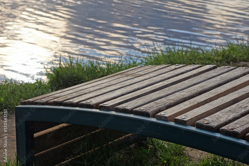 one empty brown wooden bench stands in green grass by a earth in a park on the shore of the lake with gray water