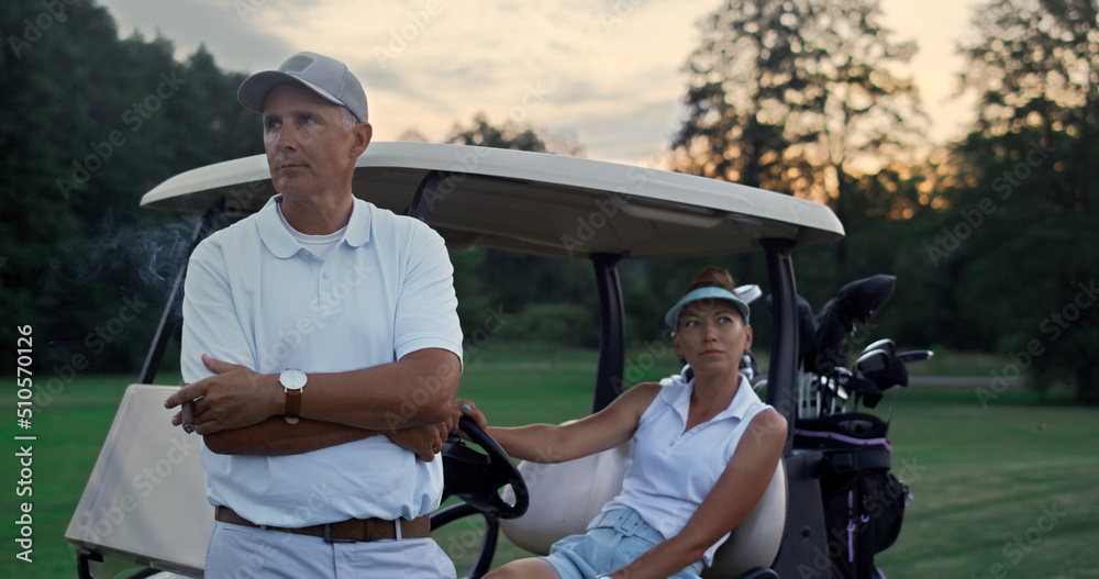Wealthy golf players resting at golf cart. Sport couple enjoy summer weekend.
