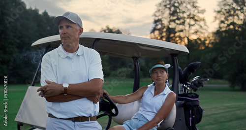 Wealthy golf players resting at golf cart. Sport couple enjoy summer weekend.