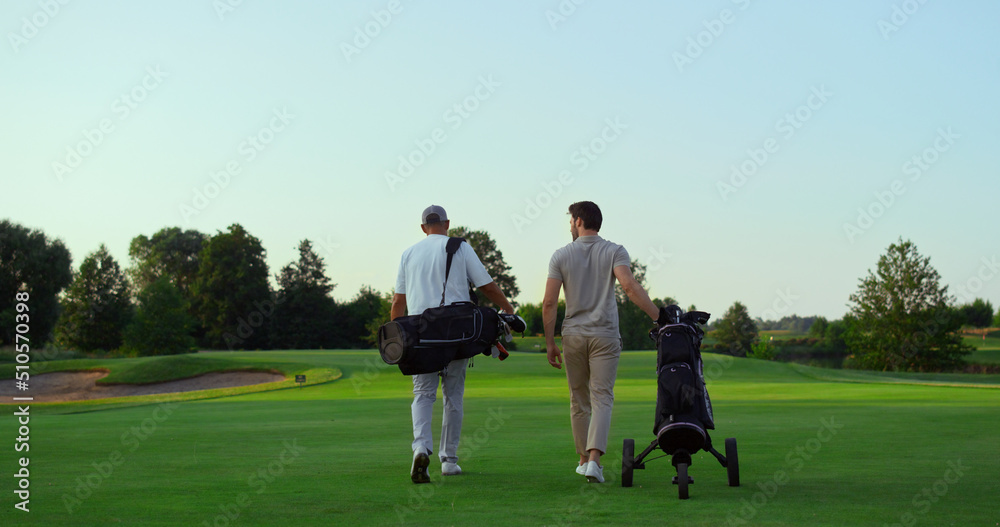 Rich golf players walking on field. Two active sportsmen carry equipment clubs.