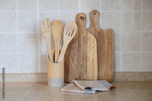 Different kitchen utensil made of wood in a holder, cutting board and a blanket on marble tabletop. Copy space for text, close up, wall with beige facing tile background. photo
