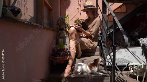 writting notes sitting on wooden stairs in contryside style yard with small dog. Relaxed summer day mood. Woman in linen natural dress and straw dress. Small dog lies at the feet. Video footage photo