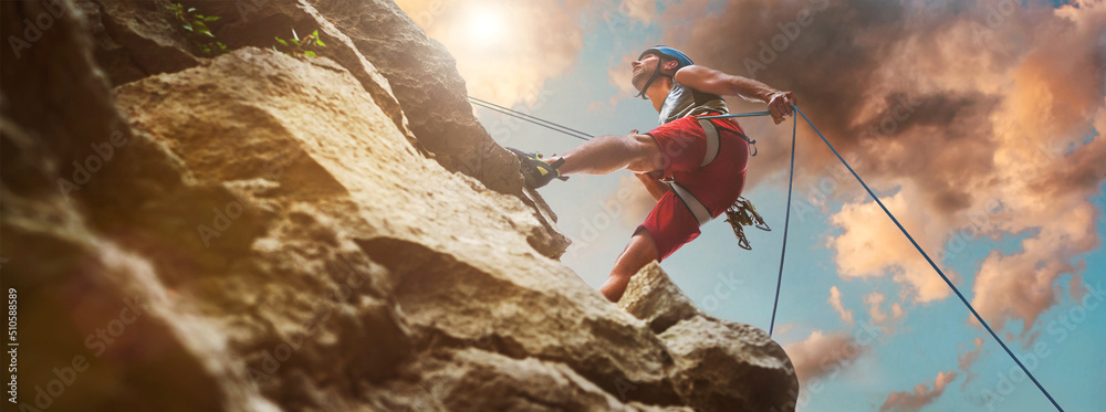 Muscular climber man in protective helmet abseiling from cliff rock wall using rope Belay device and climbing harness on evening sunset sky background. Active extreme sports time spending concept. - obrazy, fototapety, plakaty 