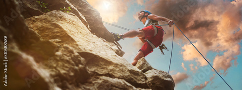 Obraz na płótnie Muscular climber man in protective helmet abseiling from cliff rock wall using rope Belay device and climbing harness on evening sunset sky background