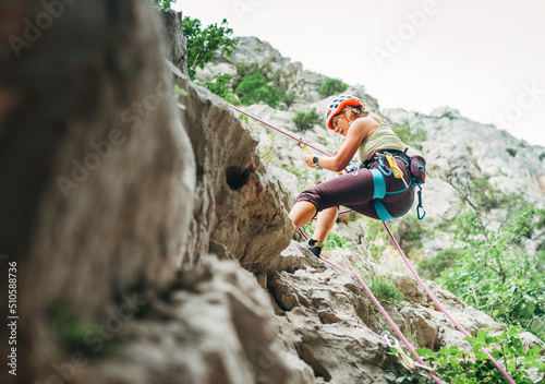 Active climber woman in protective helmet abseiling from cliff rock wall using rope with belay device and climbing harness. Active extreme sports time spending concept.