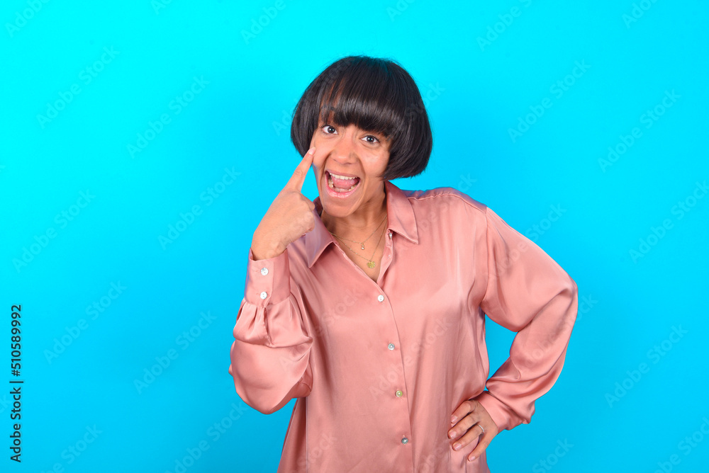 young brunette woman wearing pink silk shirt over blue background, looking, observing, keeping an eye on an object in front, or watching out for something. young brunette woman wearing pink silk shirt over blue background, looking, observing, keeping an eye on an object in front, or watching out for something.