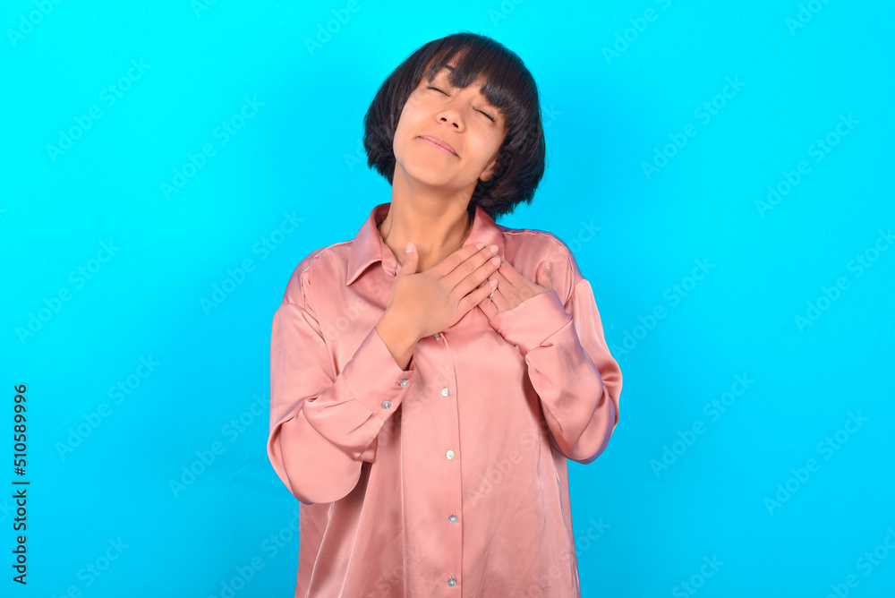 young brunette woman wearing pink silk shirt over b closes eyes and keeps hands on chest near heart, expresses sincere emotions, being kind hearted and honest. Body language and real feelings concept. young brunette woman wearing pink silk shirt over b closes eyes and keeps hands on chest near heart, expresses sincere emotions, being kind hearted and honest. Body language and real feelings concept.