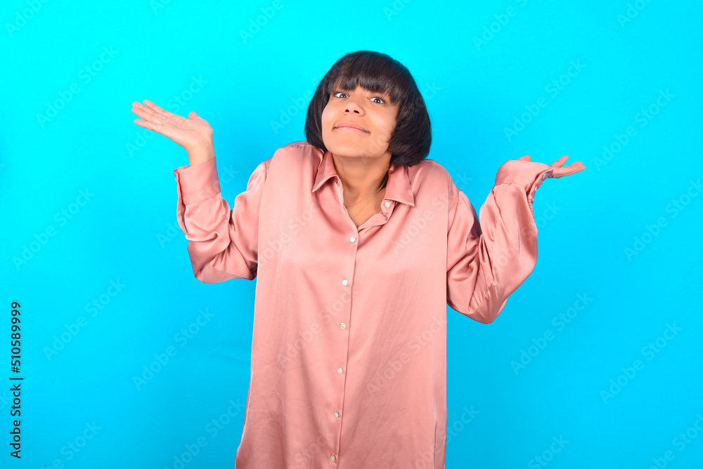 Maybe yes or no. Doubtful young brunette woman wearing pink silk shirt over blue background shrugs shoulders in bewilderment, tries to make decision puzzled what he wants. Hesitation and uncertainty. Maybe yes or no. Doubtful young brunette woman wearing pink silk shirt over blue background shrugs shoulders in bewilderment, tries to make decision puzzled what he wants. Hesitation and uncertainty.