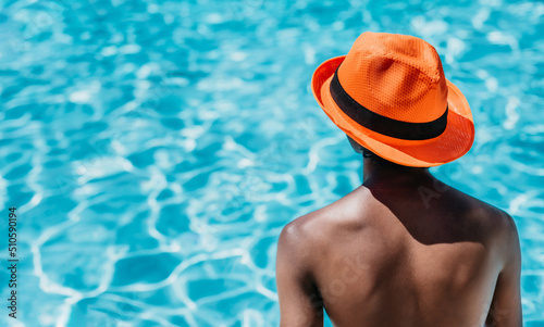 a boy of indian origin looks sideways with his back to the blue water of a swimming pool. he is wearing a striking orange hat. concept vacations and diversity. horizontal format © Karlos Garciapons