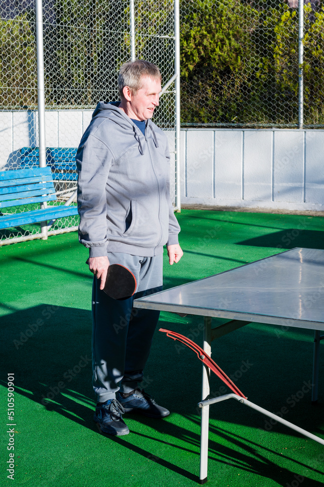 An elderly, gray-haired retired man plays table tennis ping pong on an outdoor sports field on a sunny day. Motion blur on the ball. The concept of an active lifestyle of elderly pensioners An elderly, gray-haired retired man plays table tennis ping pong on an outdoor sports field on a sunny day. Motion blur on the ball. The concept of an active lifestyle of elderly pensioners