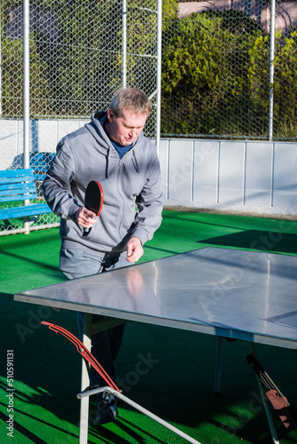 An elderly, gray-haired retired man plays table tennis ping pong on an outdoor sports field on a sunny day. Motion blur on the ball. The concept of an active lifestyle of elderly pensioners