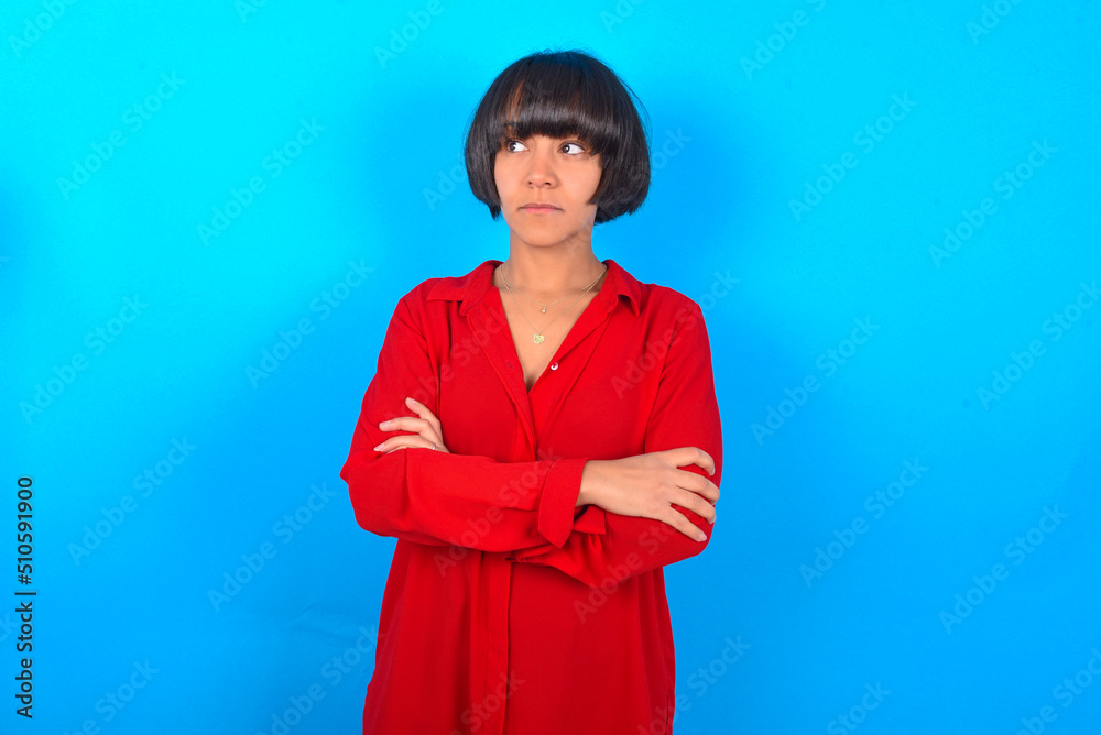 Charming thoughtful young brunette woman with short hair wearing red shirt over blue background stands with arms folded concentrated somewhere with pensive expression thinks what to do Charming thoughtful young brunette woman with short hair wearing red shirt over blue background stands with arms folded concentrated somewhere with pensive expression thinks what to do