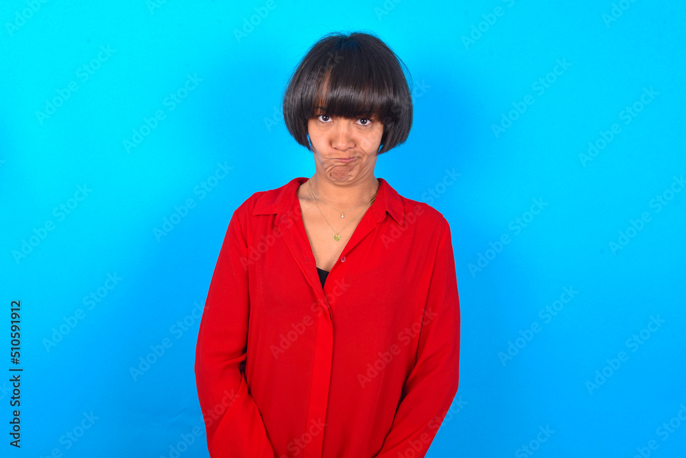 Fototapeta premium Offended dissatisfied young brunette woman with short hair wearing red shirt over blue background with moody displeased expression at camera being disappointed by something