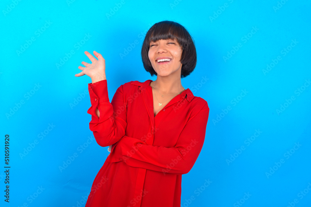 Overjoyed successful young brunette woman with short hair wearing red shirt over blue background raises palm and closes eyes in joy being entertained by friends Overjoyed successful young brunette woman with short hair wearing red shirt over blue background raises palm and closes eyes in joy being entertained by friends