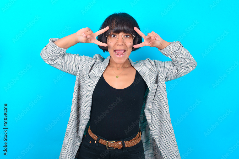Cheerful positive young businesswoman with bob haircut wearing blazer against blue wall shows v-sign near eyes open mouth Cheerful positive young businesswoman with bob haircut wearing blazer against blue wall shows v-sign near eyes open mouth