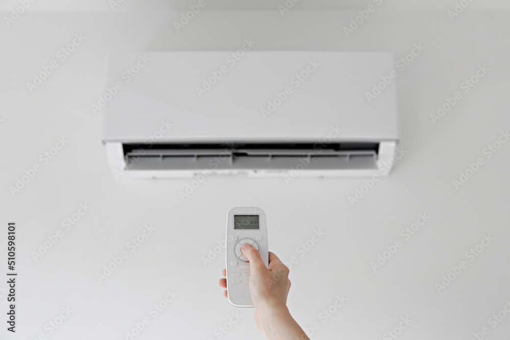 Close up shot of a woman's hand holding the remote control of newly installed white air conditioner. AC hanging on the wall working in a cooling mode set to 24 degrees celsius. Background, copy space. Close up shot of a woman's hand holding the remote control of newly installed white air conditioner. AC hanging on the wall working in a cooling mode set to 24 degrees celsius. Background, copy space.