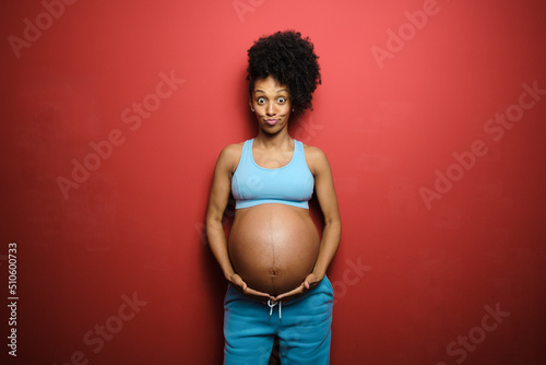 Fun portrait of surprised healthy expressive black pregnant woman on fitness sportswear against red background.