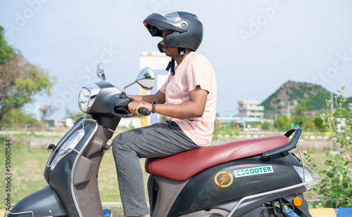 Fotografia, Obraz Man driving Ev or electric scooter by wearing helmet on road - concept of safety, technology and lifestyle