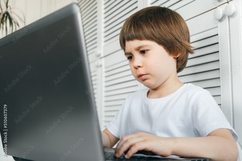5 year boy sits at the table, uses laptop, looking very close and long time at the screen. Unhappy child does homework lesson. Hard question. Damage to vision. Home distance online education concept