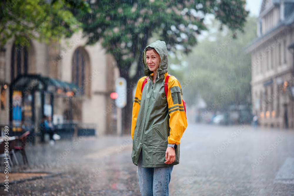 Fototapeta premium Young woman in green yellow raincoat stands happily on street under the rain