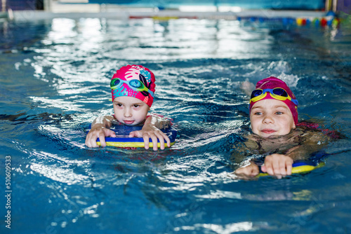 Children learn to swim with board in pool under guidance of coach. Active kids are playing in water.