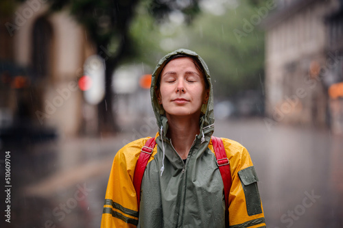 Young woman in green yellow raincoat stands happily on street under the rain