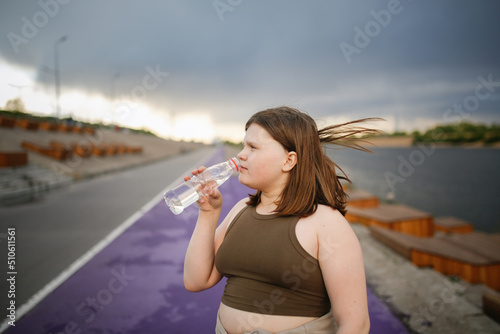 Overweight European teenage girl in tracksuit drinks water from bottle while jogging along on concrete embankment, Sports and overweight teenagers