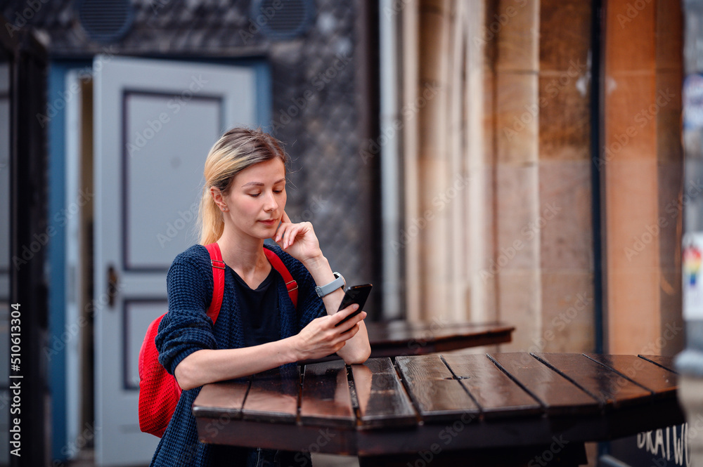 Fototapeta premium Young blonde woman in autumn-spring outwear and red backpack stands near high wooden table
