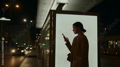 A young woman stands at a bus stop at night and uses a smartphone