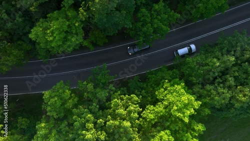 The view from the drone to the road through the forest against the background of the setting sun. Aerial view of cars driving through the forest. Forest road from the bird's eye view.
