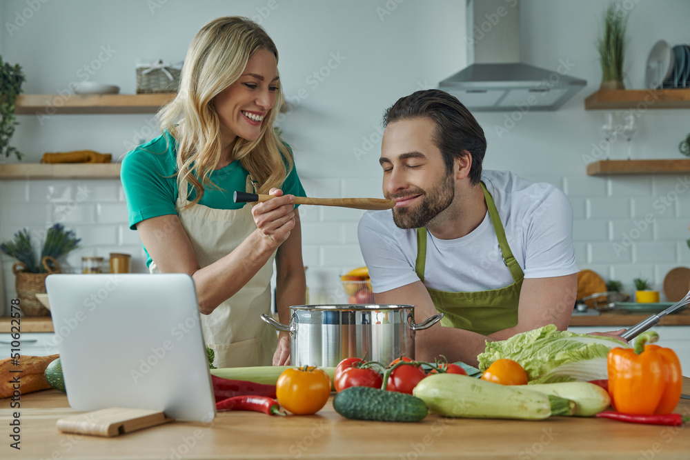 Young woman giving her husband to taste a meal while both standing at the kitchen Young woman giving her husband to taste a meal while both standing at the kitchen