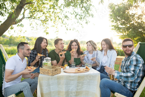 Group of young friends enjoying a picnic sitting outdoors fresh air spending weekend sunny day drinking beverage clinking cups  soft focus