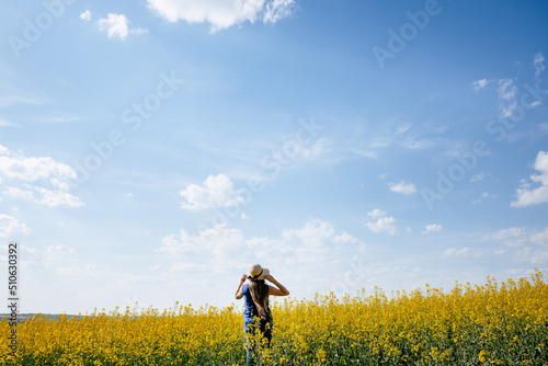 Woman wearing hat stands outside in a yellow summer field with blue sky and clouds, concept of freedom