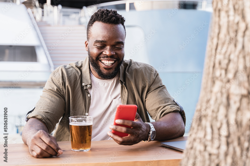 Happy African man drinking beer at brewery bar using mobile phone outdoor with luxury yacht in background - Focus on face Happy African man drinking beer at brewery bar using mobile phone outdoor with luxury yacht in background - Focus on face